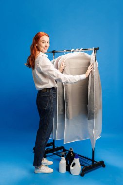 Çamaşırhane çalışanı katlanmış kıyafetleri kontrol ediyor, mavi arka planda giysi torbaları ve deterjanlarla rafların yanında duruyor. Meslek anlayışı, otel personeli, temizlik hizmeti, oda servisi.