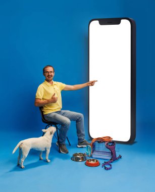 Sarı tişörtlü neşeli bir adam köpeğiyle birlikte merhaba diyor ve mavi arka planda beyaz ekranlı büyük bir cep telefonunu işaret ediyor. Evcil hayvan kavramı, hayvan bakımı, online alışveriş