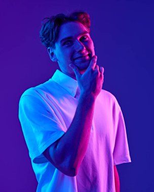 Beyaz gömlekli bir gencin portresi, neon ışıkta mavi arkaplana karşı komik mim yüzünü ifade ediyor. Gençlik kavramı, insani duygular, moda, olumlu duygular