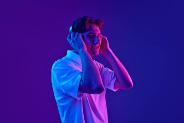 Mavi kulaklık takmış, beyaz polo tişörtlü gülümseyen ve neon ışıkta mor arka planda müzik dinleyen genç adam. Gençlik kavramı, insani duygular, moda