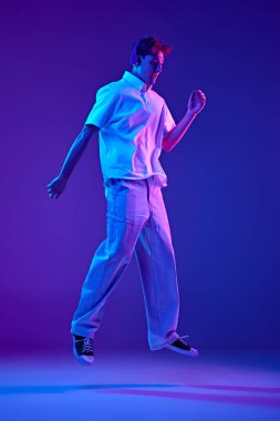 Beyaz kıyafetli duygusal bir gencin tam boy görüntüsü kulaklıkla müzik dinlerken ve neon ışıkta mavi arkaplanda dans ederken. Gençlik kavramı, insani duygular, moda