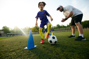 Erkek spor koçu genç oyuncuya top sürme taliminde rehberlik ediyor, cesaret veriyor ve yorum yapıyor. Odaklan ve konsantre ol. Spor, çocukluk, eğitim, başarı, aktif yaşam tarzı kavramı