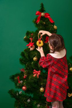 Güzel küçük bir kız, kırmızı kareli elbiseli bir çocuk ve Noel ağacını çöreklerle süsleyen, yeşil stüdyonun arka planında geçen bir çocuk. Kış tatili, Noel, Yeni Yıl kavramı