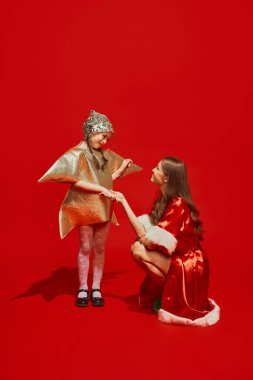 Yıldız şeklinde kostüm giymiş küçük bir kız çocuğu kırmızı stüdyo arka planında festival kostümü giymiş genç bir kadınla konuşuyor. Kış tatili kavramı, Noel, yeni yıl, kutlama, mutluluk