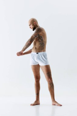Formda, rahatlatıcı, dövmeli kaslı, beyaz atölye arka planında beyaz iç çamaşırlarıyla üstsüz poz veren genç bir adamın tam boy görüntüsü. Erkek güzelliği, vücut ve sağlık hizmetleri, spor