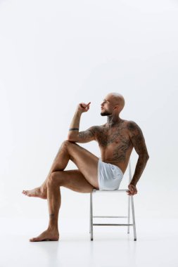 Yakışıklı, kel bir adamın yan görünüşü, beyaz iç çamaşırlarıyla sandalyede poz veren, atletik vücudu ve beyaz arka planda izole edilmiş dövmeleri gösteren bir model. Güzellik, vücut ve sağlık hizmetleri kavramı,