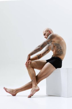 Siyah iç çamaşırlı, küpsüz oturan yakışıklı genç adam beyaz stüdyo arka planında izole edilmiş güçlü ve atletik bir vücut gösteriyor. Erkek güzelliği, vücut ve sağlık hizmetleri, spor