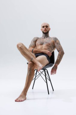 Sandalyeye oturan yakışıklı genç kel adamın portresi, iç çamaşırlarıyla üstsüz poz veriyor, beyaz stüdyo arka planında izole edilmiş kaslı vücudu gösteriyor. Erkek güzelliği, vücut ve sağlık hizmetleri, spor