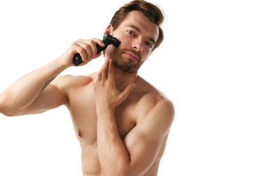 Tişörtsüz yakışıklı adam sakal tıraşı kullanıyor, beyaz arka planda izole kılları dikkatlice şekillendiriyor. Çağdaş erkek bakımı. Erkek güzelliği, cilt bakımı, kozmetik, kozmetik