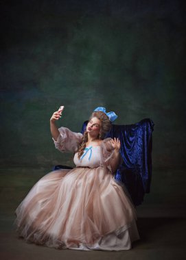 Dönem elbisesi içinde güzel bir genç kadının portresi mavi sandalyede oturmuş ve yeşil arka planda cep telefonuyla selfie çekiyordu. Tarih kavramı, Rokoko sanatı, çağların karşılaştırması