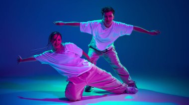 Genç adam ve kız gündelik kıyafetlerle dans ediyor, neon ışıkta hip-hop yapıyor, mor stüdyoda arka planda hip-hop yapıyor. Çağdaş dans, hobi, gençlik kavramı