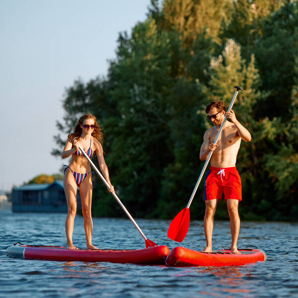 Essence of summer fun. Young man and woman in swimwear standing on paddle board in the middle of the river, with greenery on background. Concept of sport, summer, active lifestyle, hobby, leisure time — Stock Image Essence Summer Fun Young Man Woman Swimwear Standing Paddle Board Royalty Free Stock Photos