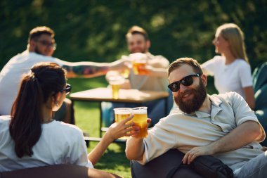 - Şerefe. Genç gülümseyen erkekler ve kadınlar, arkadaşlar kutlama yapıyor, sıcak yaz gününde dışarıda parti veriyor, neşeli bir şekilde konuşuyor ve bira içiyor. Yaz kavramı, rahatlık, arkadaşlık, toplantı, rahatlama.