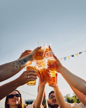 Bir grup insan, arkadaşlar piknikte toplanıyor, bardakları birayla kaldırıyor, açık mavi gökyüzünün altında şıngırdıyor. Geleneksel festival. Yaz kavramı, eğlence, arkadaşlık, toplantı, eğlence, rahatlama.