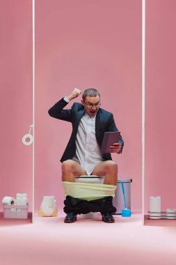 Gol! Resmi giyinmiş duygusal bir adam pantolonuyla tuvalette oturur ve tabletle futbol maçı izler ve pembe arka planda bağırır. Spor, oyun, maç etkinliği, insan duyguları kavramı