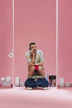 Komik adam tuvalette pantolonunu indirmiş ve cep telefonuyla pembe arka planda konuşuyor. Spor, oyun, maç etkinliği, insan duyguları kavramı