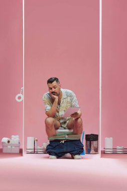Renkli gömlekli genç adam sıkılmış yüzüyle tuvalette oturuyor ve pembe arka planda tablete bakıyor. Spor, oyun, maç etkinliği, insan duyguları, televizyon programı kavramı