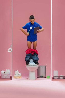 Genç bir çocuk, mavi tişörtlü bir çocuk tuvalette duruyor, elinde tuvalet fırçası ve tablet var, pembe arka planda maç çevirisi izliyor. Spor, oyun, maç etkinliği, insan duyguları kavramı