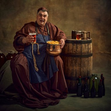 Cheerful man, medieval monk, holding pint of ale, beer and pot of chips, joyfully sitting against vintage background. Rustic monastery settings. Concept of comparison of eras, brewery, traditions clipart