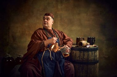 Ortaçağ keşişi giysileri içinde çaydanlıktan bira dolduran, loş ışıklı mahzende oturan, üzerinde tahta fıçı ve bira bardağı olan bir keşiş. Oktoberfest. Devir, bira fabrikası, gelenek karşılaştırması kavramı