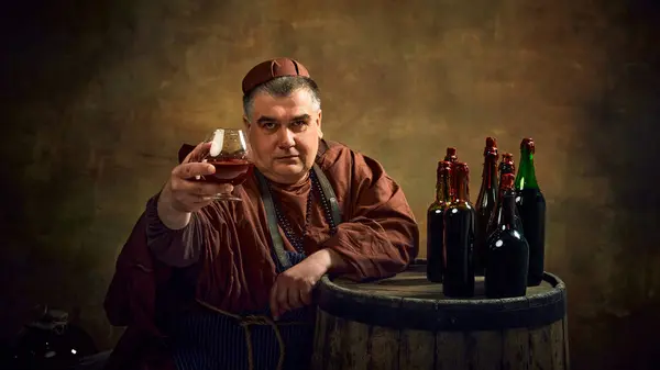 stock image Man, medieval monk in robe raising glass filled with brown liquid, beer, ale, cognac against vintage background. Traditions of abbey brewing. Concept of comparison of eras, history, brewery