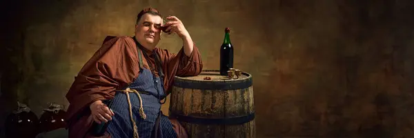 Stock image Hangover. Medieval man, monk in traditional attire leaning glass with beverage to head, leaning on wooden barrel with bottle. Concept of comparison of eras, history, brewery, traditions. Banner