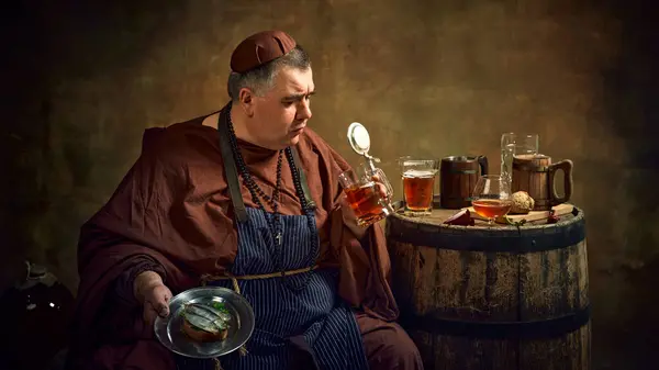 stock image Medieval man, monk, with ale mug and plate of fish sandwich degustating craft beer against vintage background. Brewing and abbey ale production. Concept of comparison of eras, history, traditions