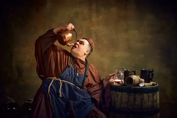 stock image Medieval monk in period clothes drinking beer from kettle, sitting in dimly lit cellar with wooden barrel and beer mug on top. Oktoberfest. Concept of comparison of eras, history, brewery, traditions