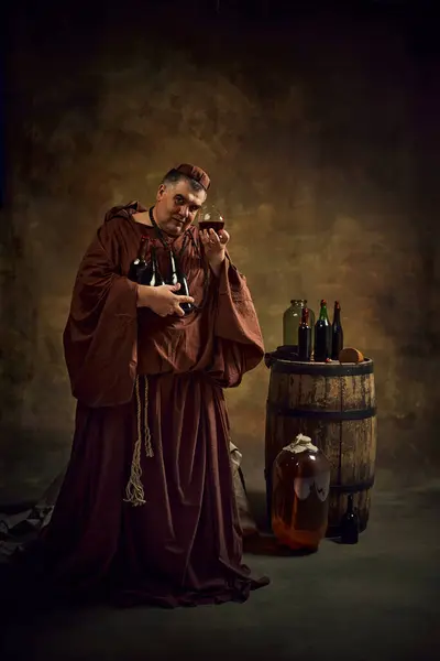 stock image Medieval monk in period attire holding glass with cognac, beer, ale again vintage cellar with wooden barrel and bottles in top. Abbey production. Concept of comparison of eras, brewery, traditions