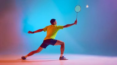 Atletik genç adam, badminton oyuncusu, hareket eğitiminde, neon ışıkta, gradyan mavi pembe arka plana karşı maça hazırlanıyor. Spor anlayışı, rekabet, aktif ve sağlıklı yaşam tarzı