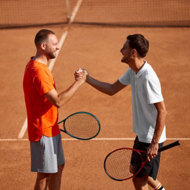 İki gülümseyen adam, kil sahadaki tenis oyuncuları maç öncesi tokalaşıyorlar, takım çalışması ve iyi bir eğitime hazır olduklarını gösteriyorlar. Spor anlayışı, rekabet, aktif ve sağlıklı yaşam tarzı