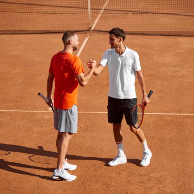 Genç adam, tenis oyuncusu antrenörüyle açık hava kil kortunda buluşuyor, erkekler yoğun antrenmandan önce tokalaşıyorlar. Spor anlayışı, rekabet, aktif ve sağlıklı yaşam tarzı