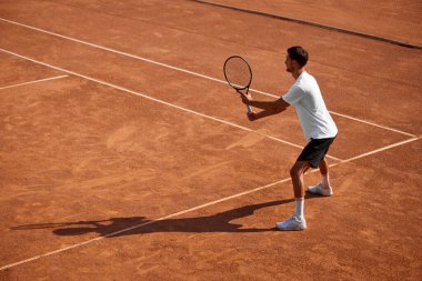 Açık hava kil sahada oynanan yoğun tenis maçında beyaz tişörtlü ve siyah tişörtlü odaklanmış genç adam topu almaya hazır olduğunu gösteriyor. Spor anlayışı, rekabet, aktif ve sağlıklı yaşam tarzı