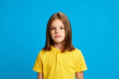 9 yaşında, kahverengi saçlı, pembe polo tişörtlü, sakin yüzüyle kameraya bakan, mavi stüdyo arka planında poz veren bir kızın portresi. Çocukluk kavramı, çocuk duyguları, yaşam tarzı