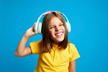 Sarı polo tişörtlü 9 yaşındaki gülümseyen kızın mavi stüdyo arka planında müzik dinlemek için kulaklık kullanmasının portresi. Çocukluk kavramı, çocuk duyguları, yaşam tarzı. aygıtlar
