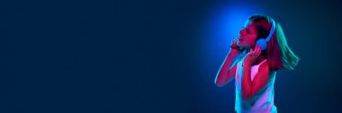Kulaklıkla müzik dinleyen genç bir kız mavi stüdyonun arka planına karşı neon ışıkta. Eğlence ve eğlence aktivitesi. Çocukluk kavramı, çocuk duyguları, yaşam tarzı. Reklam afişi
