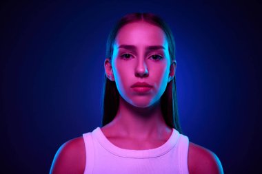 Güzel genç bir kadının portresi, özgüvenli poz veren öğrenci, neon ışıklarıyla değişen mavi stüdyo arkaplanına bakıyor. İnsan duyguları kavramı, günlük moda, yaşam tarzı, eğitim