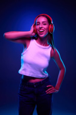 Kot pantolonlu ve beyaz bluzlu gülümseyen genç bir kadının portresi kulaklıkla müzik dinlerken neon ışıkta mavi renkli bir stüdyonun arka planına karşı. İnsani duygular, boş zaman kavramı
