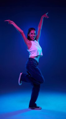 Kot pantolonlu ve beyaz bluzlu gülümseyen genç bir kadının, neon ışıklı, gradyan mavi stüdyo arka planına karşı neşeli bir şekilde poz verişinin tam boy portresi. İnsan duyguları, gençlik, moda, neşe ve eğlence kavramı