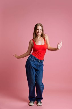 Mutluluk ve eğlence. Gündelik giysiler içinde genç güzel bir kızın portresi, kırmızı bluz ve kotlar neşeyle poz veriyor, pembe stüdyo arka planında dans ediyor. İnsan duyguları, moda, enerji, gençlik kavramı