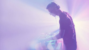 DJ dümende duruyor, dumanla çevrili, elleri neon ışıklı havada yankılanan şarkıları ustalıkla harmanlıyor. Modern müzik ve teknoloji kavramı, parti, disko, gece hayatı.
