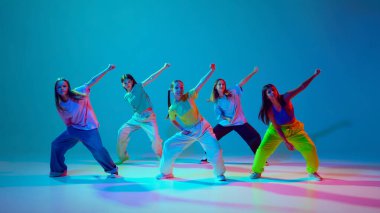 Genç kızlar dinamik performans sergiliyor, senkronize hip hop hareketlerini neon arka planda siyan mavi arka plana karşı gösteriyorlar. Modern dans stilleri, gençlik kültürü, aktif yaşam tarzı kavramı
