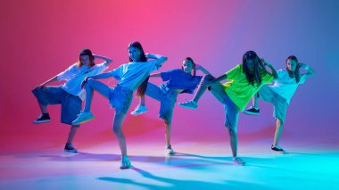 Ateşli hip hop pozları veren genç kızlar, neon ışıklarıyla değişken pembe arka plana karşı enerjik bir şekilde dans ediyorlar. Modern dans stilleri, gençlik kültürü, aktif yaşam tarzı kavramı