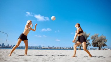 İki kız, plajda arkadaşlar açık mavi gökyüzünün altında plaj voleybolu ve arka planda şehir binası oynuyorlar. Isıtmalı plaj maçı. Spor anlayışı, yaz etkinliği, eğlence oyunları, arkadaşlık.
