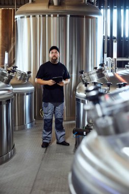 Üniformalı adam, bira fabrikası teknoloji uzmanı paslanmaz çelik tankların arasında güvenle duruyor, kaliteyi kontrol etmeye hazır. Bira, bira fabrikası, üretim, kalite kontrol kavramı