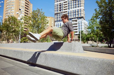 Şehir parkında antrenman yapan genç bir sporcu, elleri ve ayakları üzerinde dengede duran, taş bir bankta parkur manevrası yapan. Aktif ve sağlıklı yaşam tarzı kavramı, gençlik kültürü