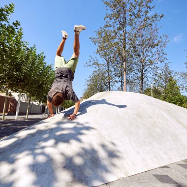 Genç sporcu kıvrımlı kentsel yapıda mükemmel amuda kalkıyor, gücü ve dengeyi açık gökyüzü ve yapraklı ağaçların arka planına karşı harmanlıyor. Aktif ve sağlıklı yaşam tarzı, gençlik kültürü kavramı