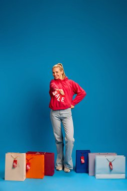 Üzerinde mavi arka planda yüzde işareti olan renkli alışveriş torbalarıyla dikilen günlük kıyafetlerle gülümseyen genç bir kadın resmi. Kara Cuma kavramı, alışveriş, satış