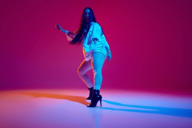 Artistik, yetenekli, büyük beden elbiseli ve topuklu ayakkabılı güzel bir genç kadın mor stüdyo arka planında yüksek topuklu dans ediyor. Hobi, eğlence, performans, yaratıcılık kavramı