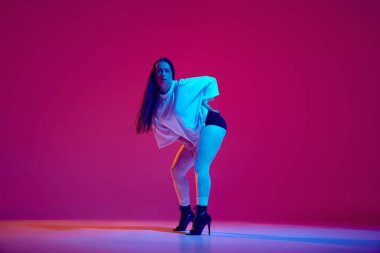 Aşırı büyük tişörtlü, siyah topuklu ayakkabılı, yüksek topuklu, eflatun renkli stüdyo arka planında dans eden şehvetli genç bir kadın. Hobi, eğlence, performans, yaratıcılık, yaşam tarzı kavramı